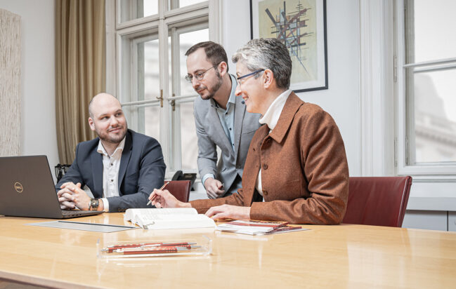 Drei Personen in Geschäftskleidung sitzen und stehen um einen Konferenztisch mit einem Laptop, einem Notebook und Dokumenten und scheinen in einem hellen Büro mit großen Fenstern und einem gerahmten Kunstwerk an der Wand über ihre Arbeit zu sprechen.